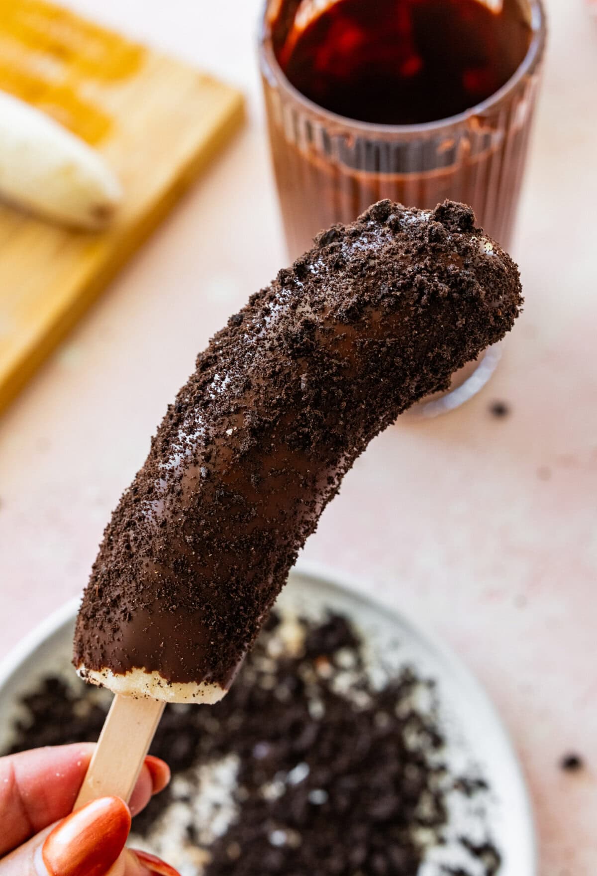 a chocolate dipped banana with oreo pieces on it, held over a plate of more oreo pieces and ingredients.