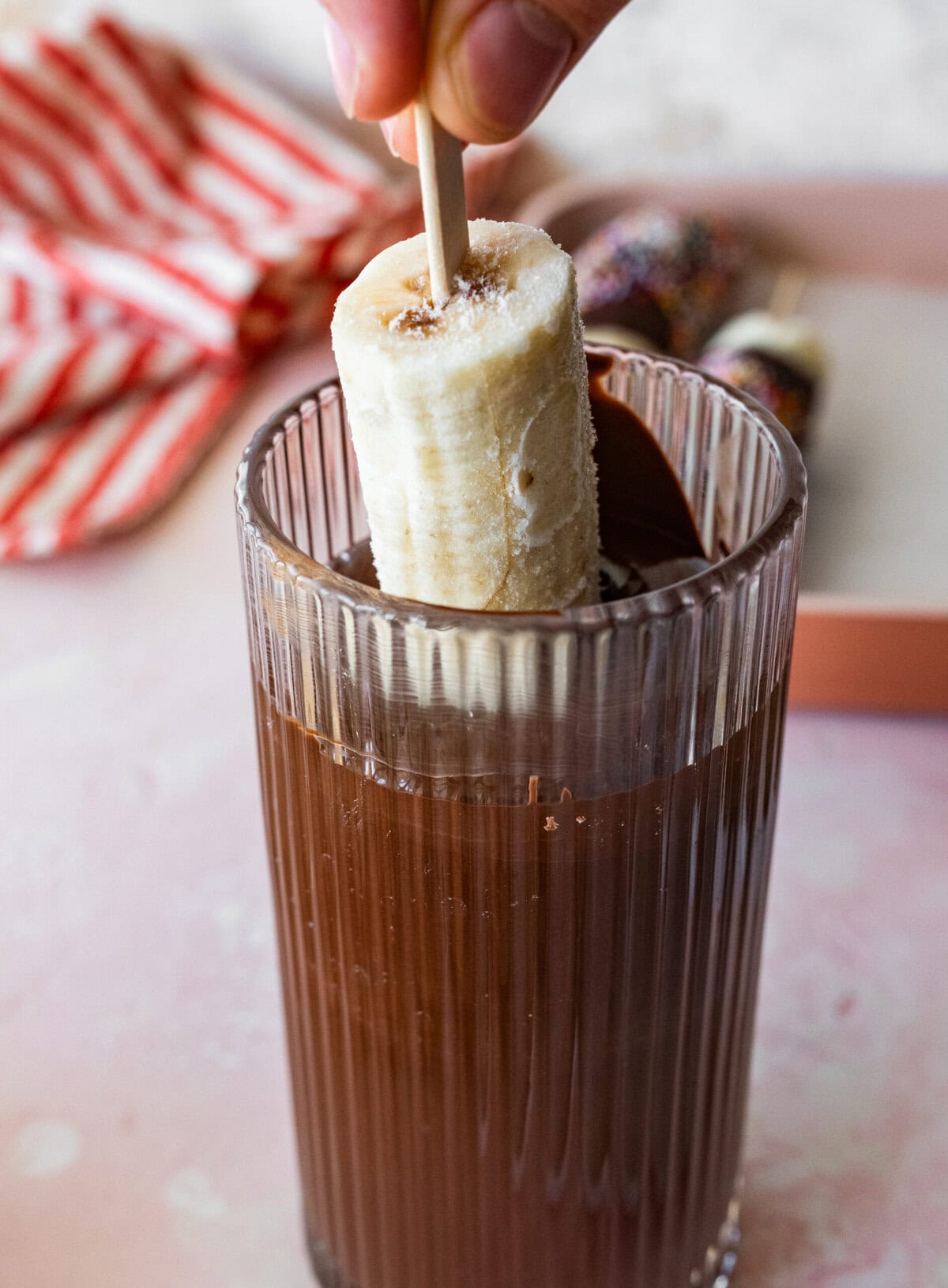 holding a banana halfway in a cup of melted chocolate.
