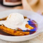 cooked bananas flambe in a white bowl with caramel sauce over the top. Ice cream on top that is slightly melted. Flame visible from the sauce.