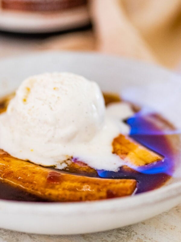cooked bananas flambe in a white bowl with caramel sauce over the top. Ice cream on top that is slightly melted. Flame visible from the sauce.