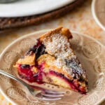 A slice of plum torte on a plate with a fork and the rest of the torte nearby.