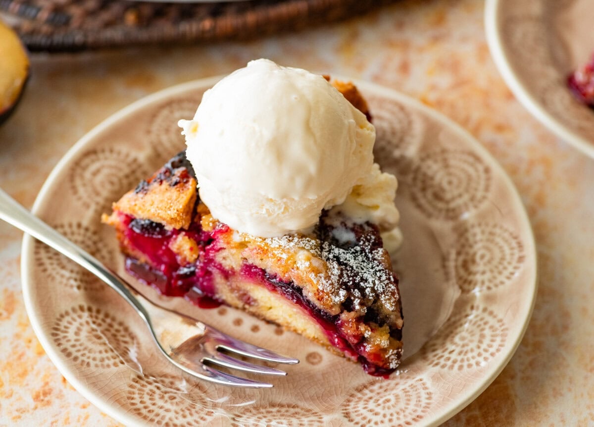 A slice of plum torte on a plate with a fork, ice cream on top, and the rest of the torte nearby.