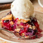 A slice of plum torte on a plate with a fork, ice cream on top, and the rest of the torte nearby.