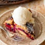 A slice of plum torte on a plate with a fork, ice cream on top, and the rest of the torte nearby.