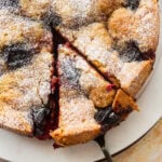 Plum torte garnished with sugar on a plate cut into slices.