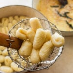 A slotted spoon removing gnocchi from the pot of water with another pot of sauce nearby.