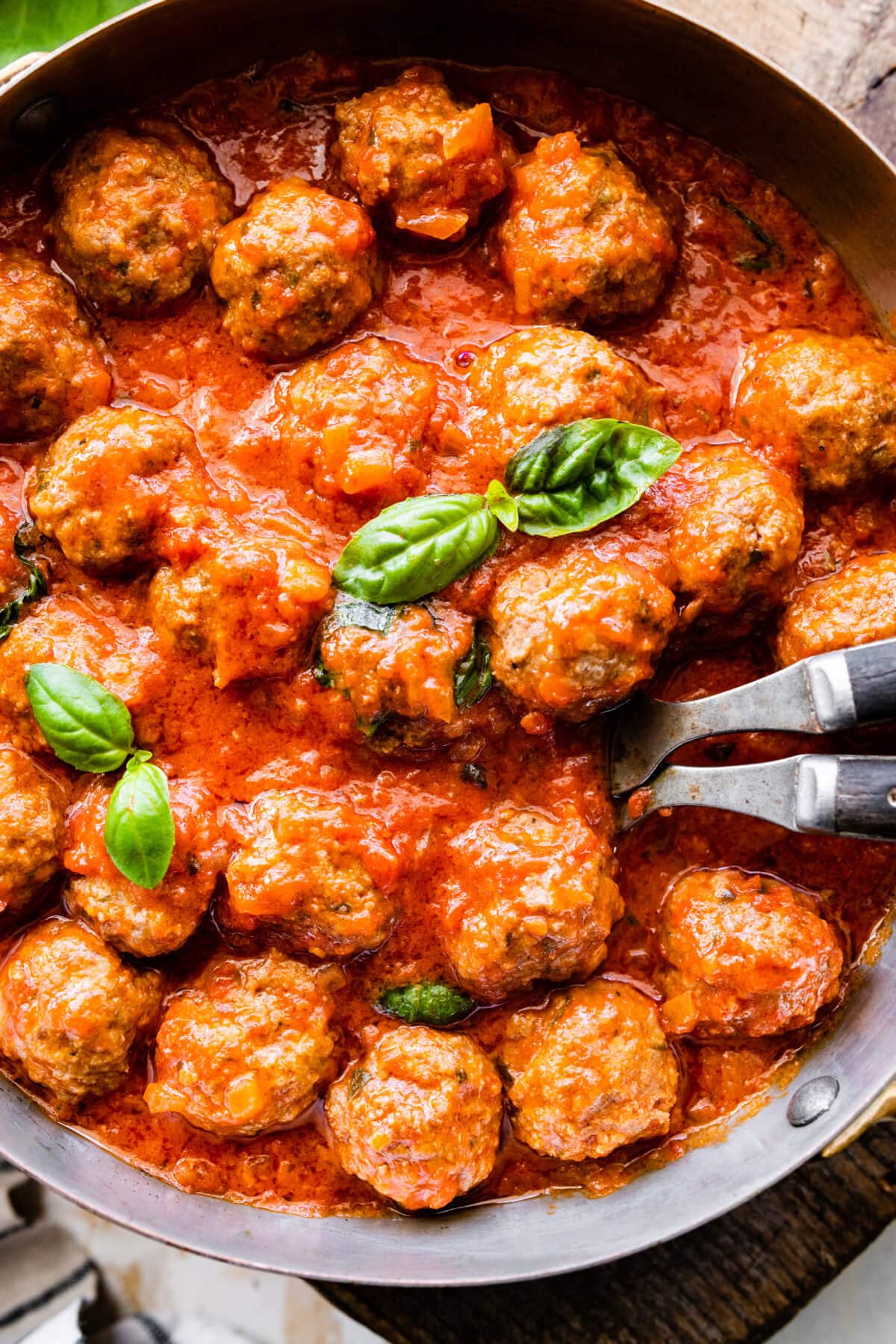 cooked meatballs in a round serving platter with serving utensils and basil on the side. Close up of the meatballs in the sauce.