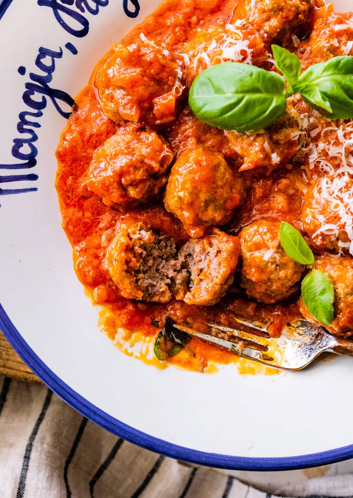 white Italian style bowl with the prepared meatballs in sauce with parmigiano cheese on top and fresh basil leaves. Tender meatballs cut in half to show tender interior.
