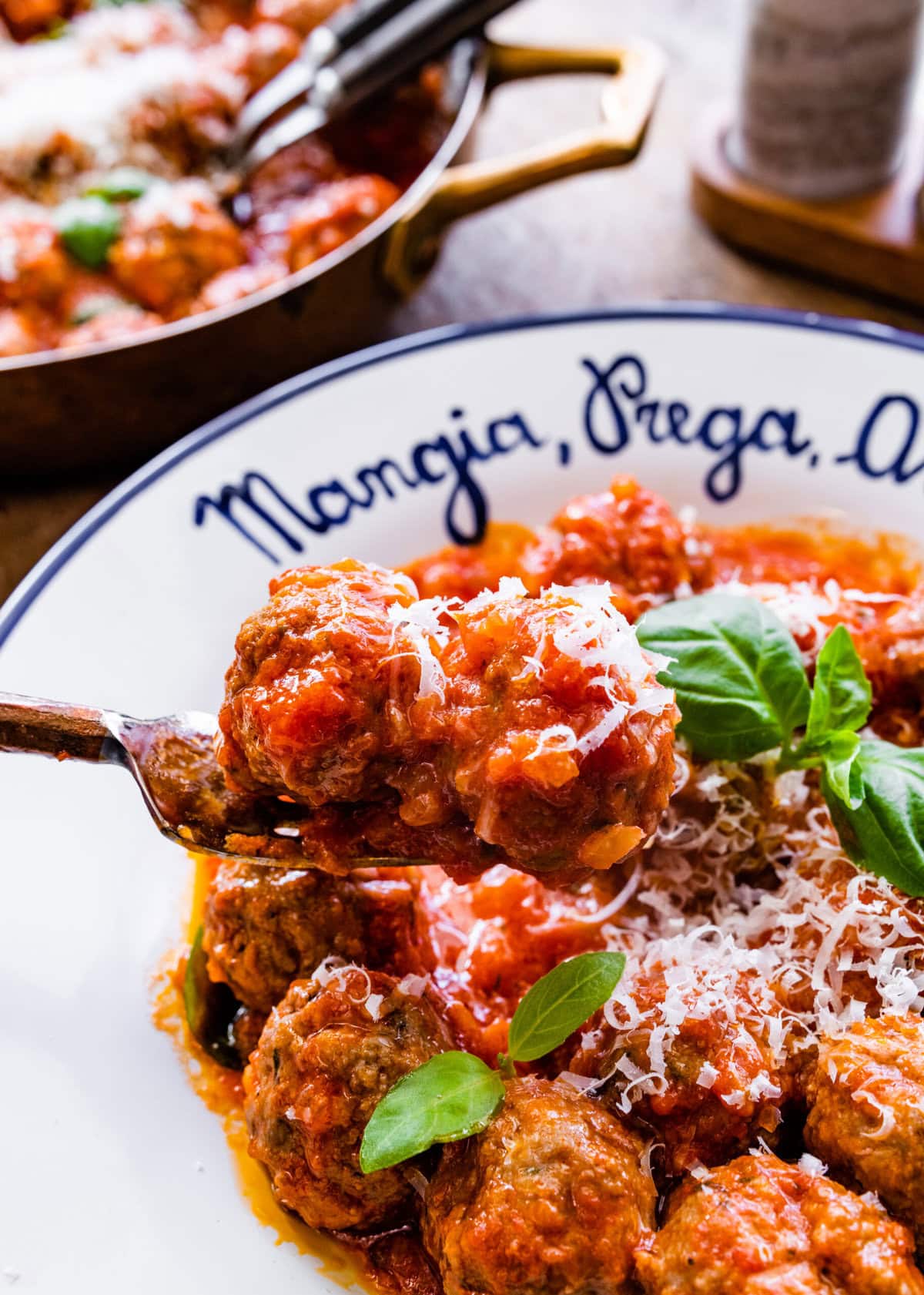 white Italian style bowl with the prepared meatballs in sauce with parmigiano cheese on top and fresh basil leaves. Fork holding up meatballs.