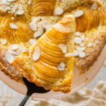 Pear frangipane tart with nuts as garnish and more ingredients and serving ware nearby with one slice being removed.