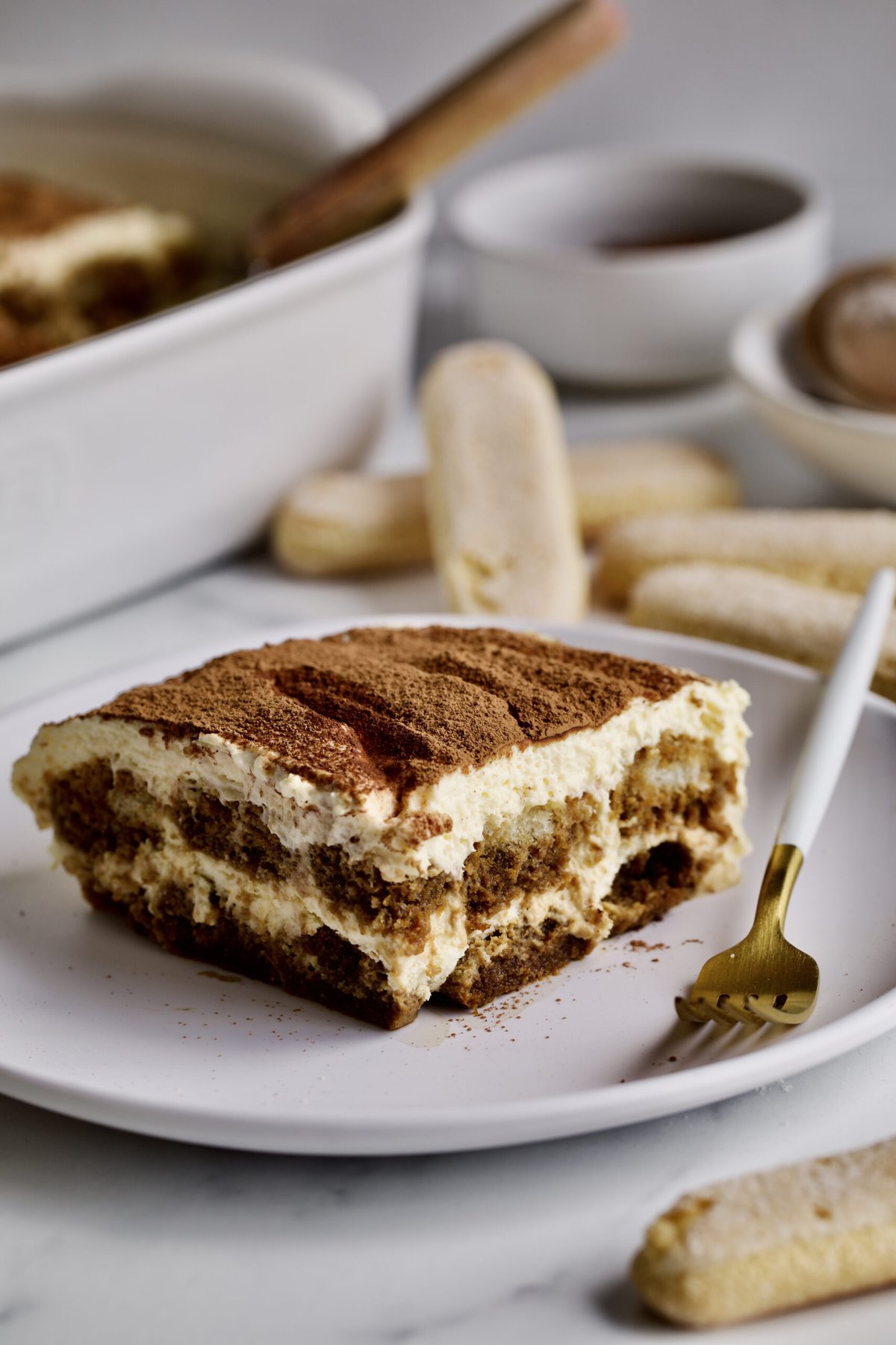 slice of tiramisu on a plate with ladyfinger cookies in the background. 