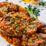 Osso buco Italian Braised Veal on a blue and white platter with gremolata sauce on top. Herbs on the side.