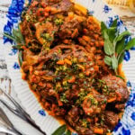 Osso buco Italian Braised Veal on a blue and white platter with gremolata sauce on top. Herbs on the side.