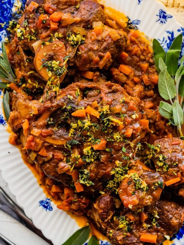 Osso buco Italian Braised Veal on a blue and white platter with gremolata sauce on top. Herbs on the side.