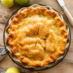 An apple pie in a pie dish with apples and a knife nearby.