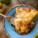 A slice of apple pie on a plate with a fork removing a bite and apples nearby.