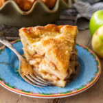 A slice of apple pie on a plate with a fork and apples nearby.