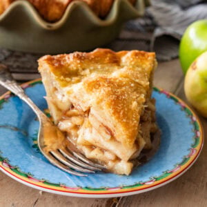A slice of apple pie on a plate with a fork and apples nearby.