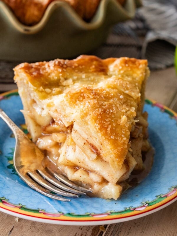 A slice of apple pie on a plate with a fork and apples nearby.