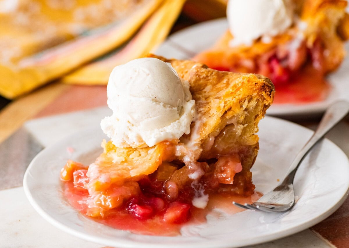 a slice of rhubarb pie on a plate, topped with ice cream with a fork and more pie nearby.
