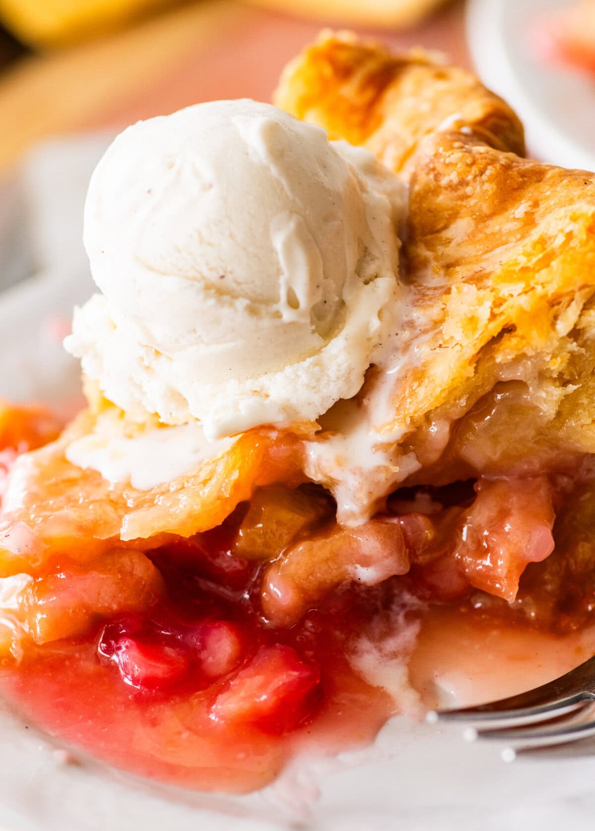 a slice of rhubarb pie on a plate, topped with ice cream with a fork nearby.