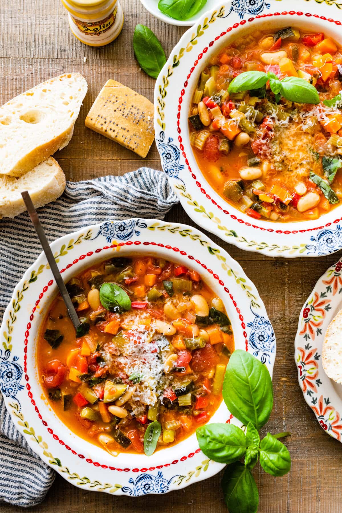 Minestrone soup in a beautiful old fashioned serving bowl with a spoon in the bowl ready to eat. Parmigiano cheese on top. Crusty bread on the side.