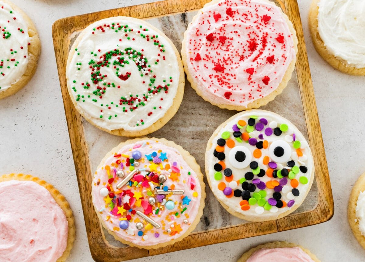 4 soft sugar cookies with frosting and different colored sprinkles on a plate with more surrounding it.