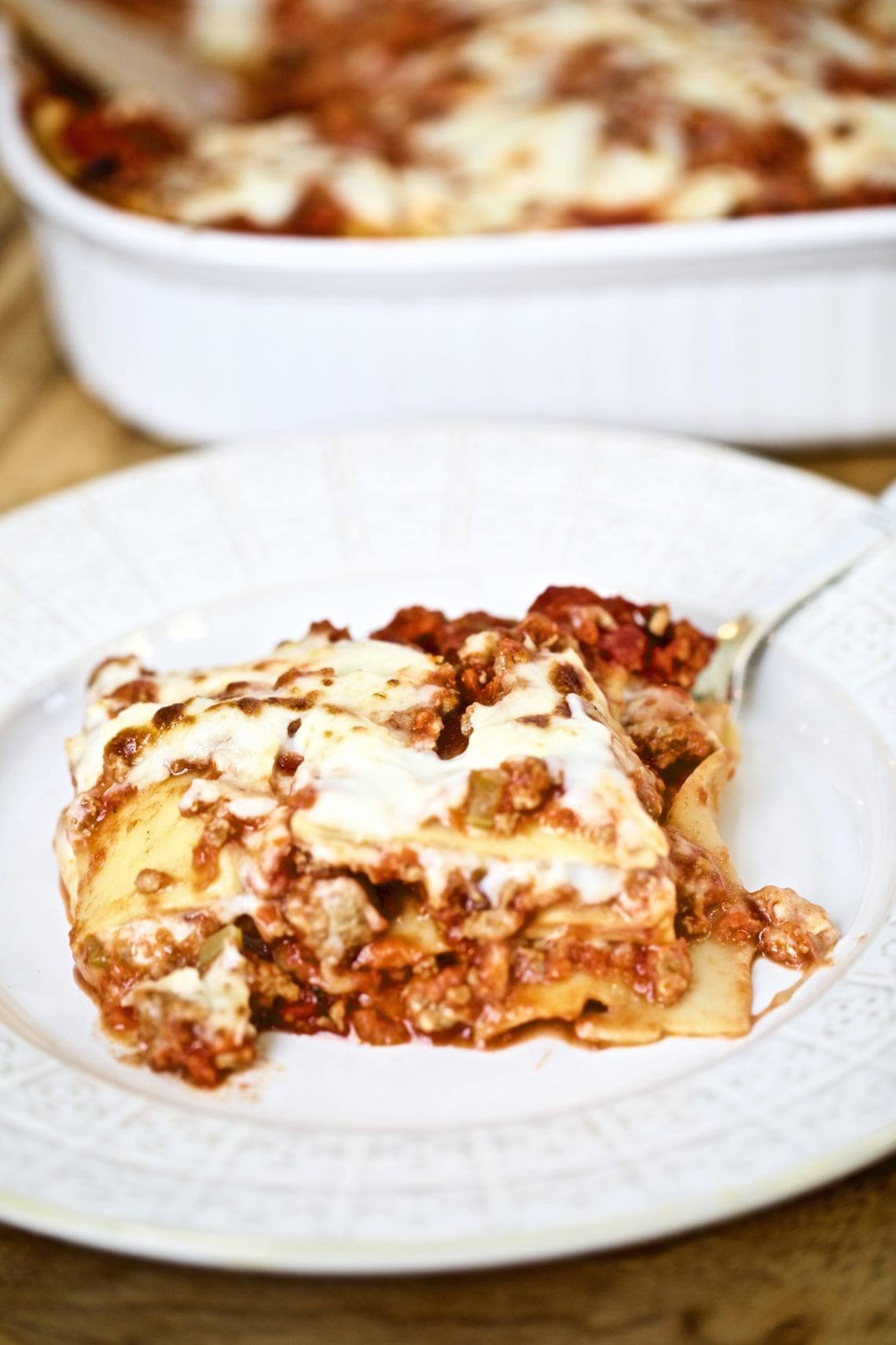 piece of square lasagna on a plate with a large lasagna pan in the background