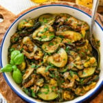 A skillet of sauteed zucchini with onions garnished with basil and parsley with a spoon resting inside and more ingredients nearby,