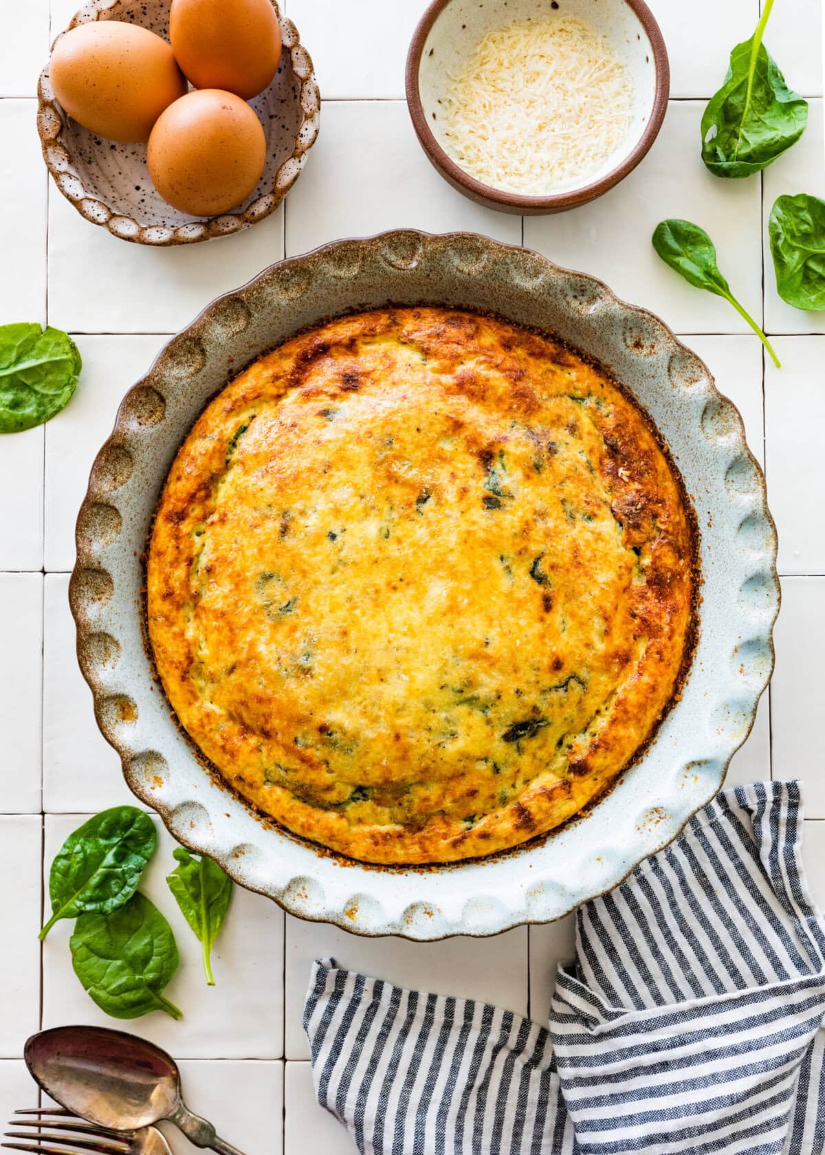Baked spinach frittata on a counter while bowls of cheese, whole eggs, and fresh herbs.
