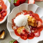 two serving bowls of strawberry rhubarb cobbler topped with ice cream with more strawberries nearby.