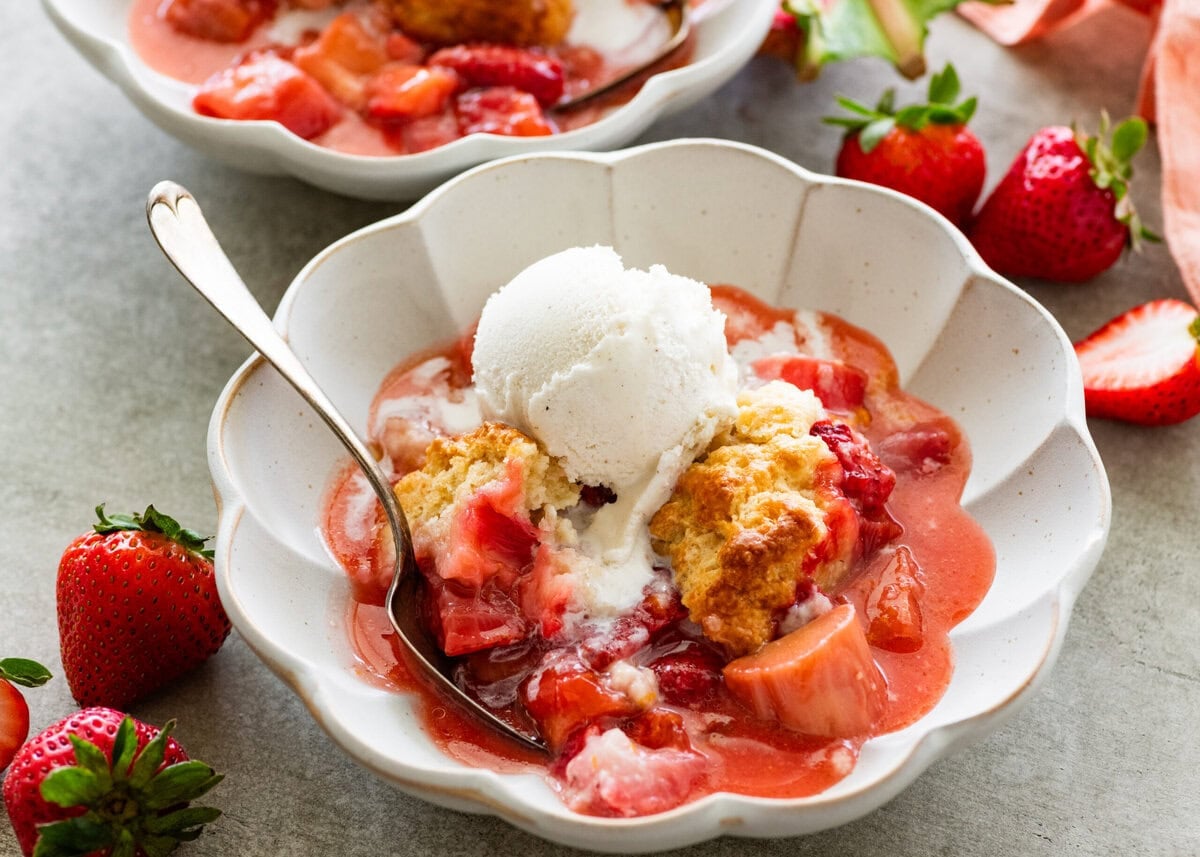 two serving bowls of strawberry rhubarb cobbler topped with ice cream with more strawberries nearby.