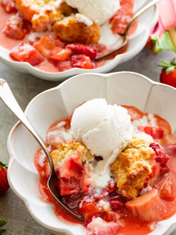 two serving bowls of strawberry rhubarb cobbler topped with ice cream with more strawberries nearby.