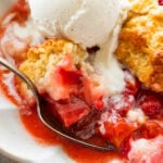 a bowl of strawberry rhubarb cobbler topped with ice cream and more strawberries nearby and a spoon holding some.