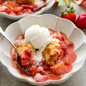 two serving bowls of strawberry rhubarb cobbler topped with ice cream with more strawberries nearby.