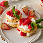 Two strawberry shortcake biscuits on a plate with more ingredients for the recipe nearby.