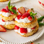 Two strawberry shortcake biscuits on a plate with more ingredients for the recipe nearby.