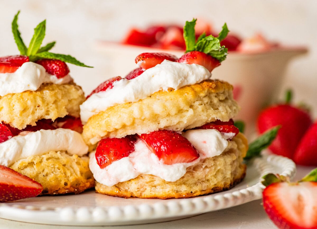 Two strawberry shortcake biscuits on a plate with more ingredients for the recipe nearby.