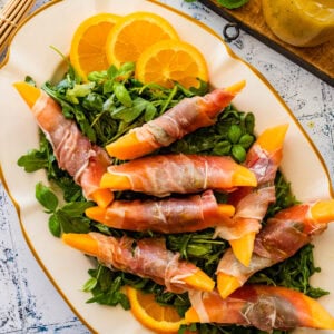 A small plate of arugula salad with sliced Prosciutto and Melon. Oranges on the side.