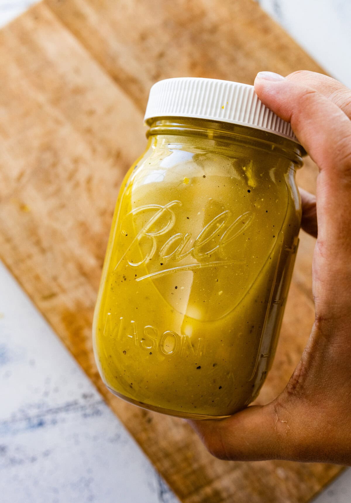 A hand holding a jar of dressing over a cutting board.