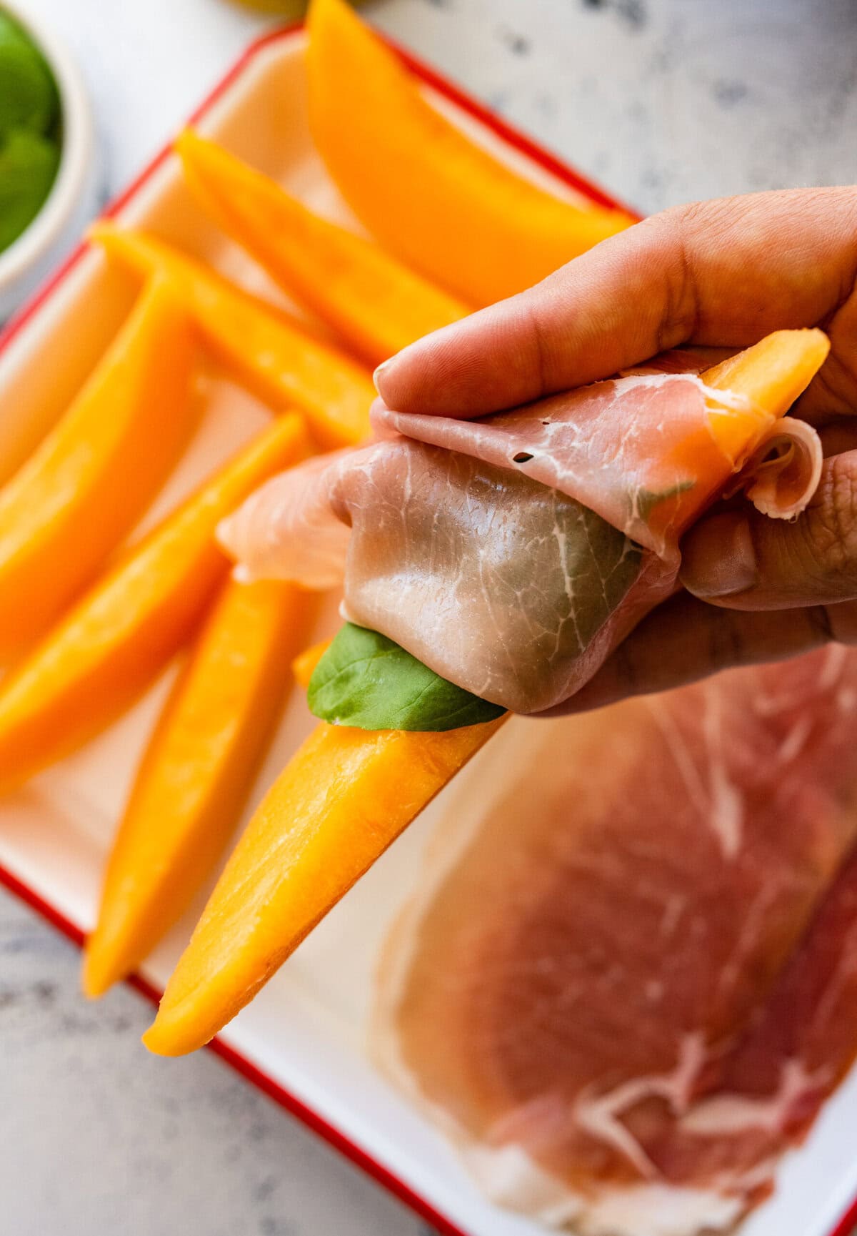 A person wrapping a piece of Prosciutto and Melon. Add basil leaves in the middle.
