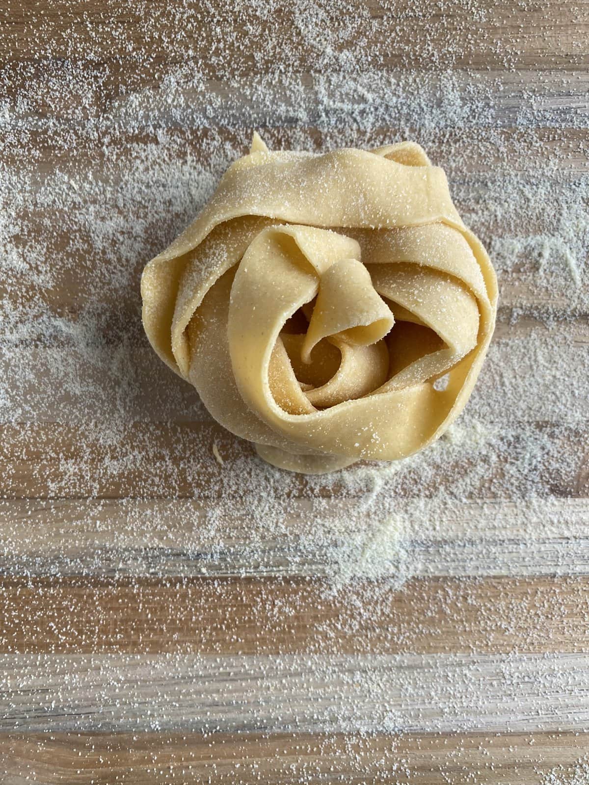 Fresh pappardelle pasta shaped into a nest on a cutting board sprinkled with semolina.