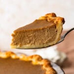 A slice of pumpkin pie being held up over the rest of the pie.
