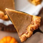 A slice of pumpkin pie being held up over the rest of the pie.