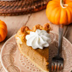 A slice of pumpkin pie on a plate, topped with whipped cream with pumpkins and the rest of the pie nearby.