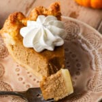 A slice of pumpkin pie on a plate, topped with whipped cream with pumpkins and the rest of the pie nearby and a bite removed with a fork.