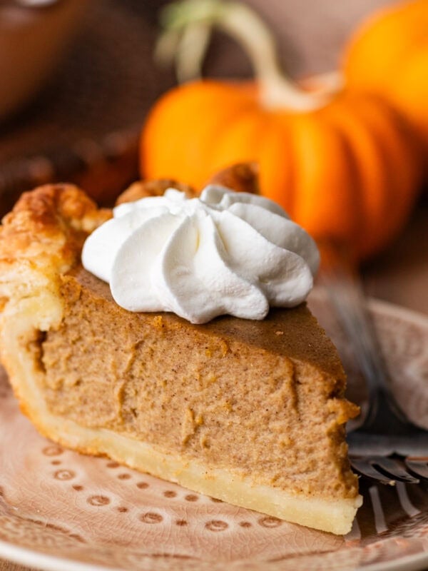 A slice of pumpkin pie on a plate, topped with whipped cream with pumpkins and the rest of the pie nearby.