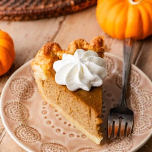 A slice of pumpkin pie on a plate, topped with whipped cream with pumpkins and the rest of the pie nearby.