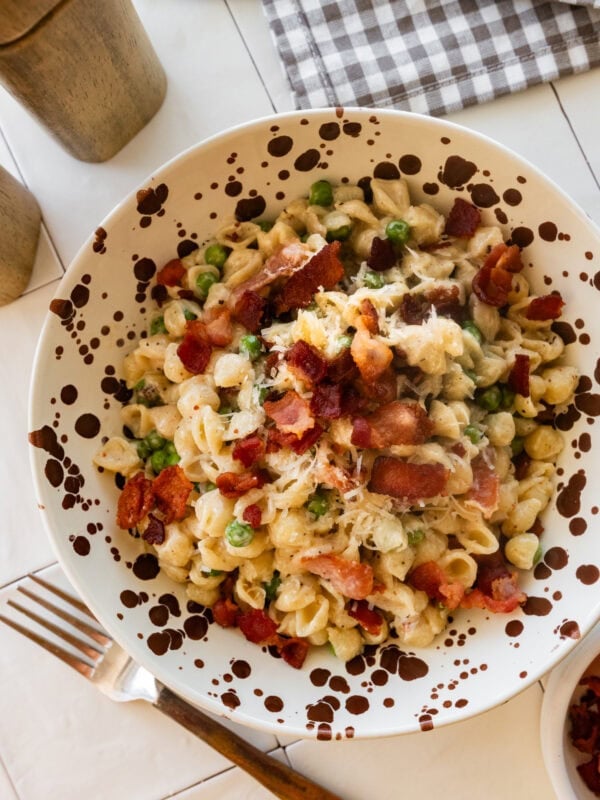 A bowl of peas and bacon pasta with a fork and more ingredients nearby.