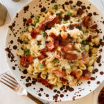 A bowl of peas and bacon pasta with a fork and more ingredients nearby.